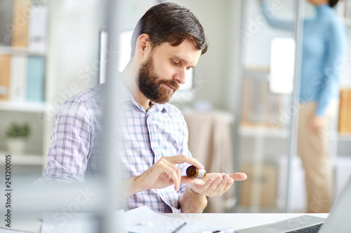 Bearded manager with headache taking pills from bottle while sitting by desk in office