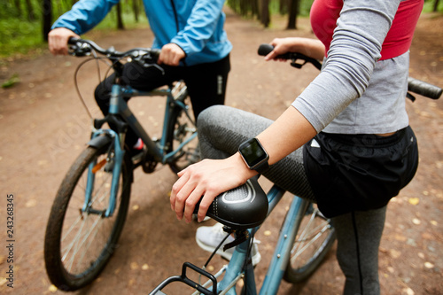 Young sportswoman with smartwatch and bicycle and her boyfriend having short break during cycle chill