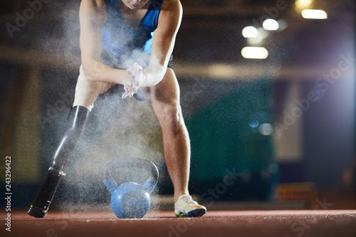 Young paralympic athlete applying talc on his hands while leaning over kettlebell at stadium