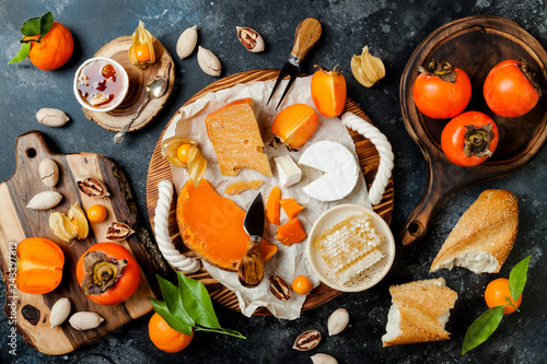 Cheese variety board or platter with cheese assortment, persimmons, honey and nuts. Black stone background. Top view, flat lay