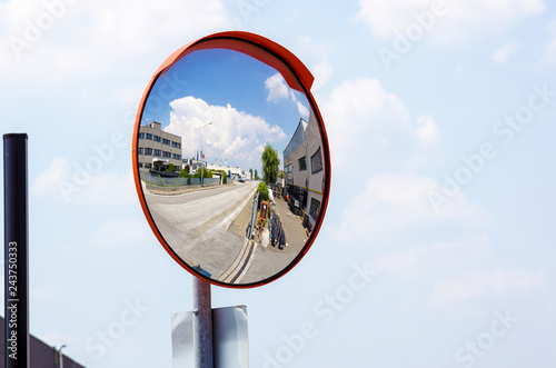 Outdoor convex safety mirror hanging on wall with reflection of an urban roadside view of cars parked along the street by residential apartment buildings.