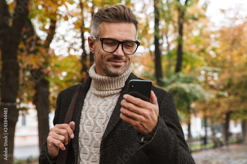 Photo of masculine man 30s wearing warm clothes walking outdoor through autumn park, and using mobile phone