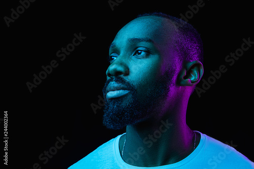 The retro wave or synth wave portrait of a young happy serious african man at studio. High Fashion male model in colorful bright neon lights posing on black background. Art design concept