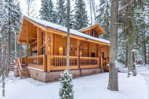 Snow-covered beautiful wooden house in the forest at dusk. Modern log cabin with large decks