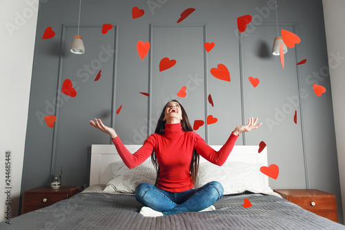 Beautiful, happy, young woman throwing red heart-shapes in the air, sitting on the bed