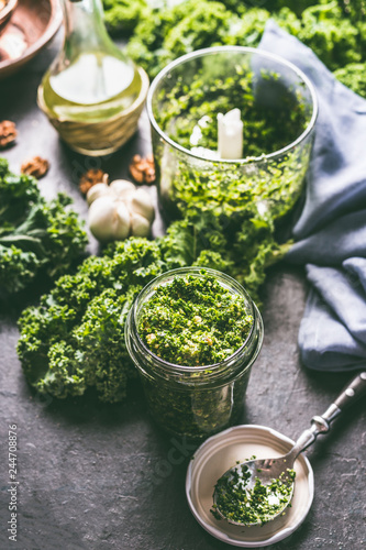 Raw kale eating recipes. Green kale pesto in glass on dark rustic kitchen table background with ingredients, top view. Kale preparation. Healthy detox vegetables . Clean eating and dieting concept.