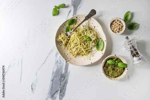 Classic italian spaghetti pasta with pesto sauce, pine nuts, olive oil and fresh basil. Served in ceramic plate with fork and ingredients above over white marble background. Flat lay, space