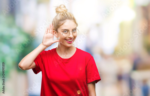 Young beautiful blonde woman wearing red t-shirt and glasses over isolated background smiling with hand over ear listening an hearing to rumor or gossip. Deafness concept.