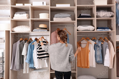 Woman choosing outfit from large wardrobe closet with stylish clothes and home stuff