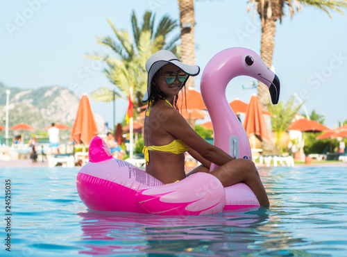 Stunning girl in bikini sits in a pool with blue water on a pink flamingo mattress