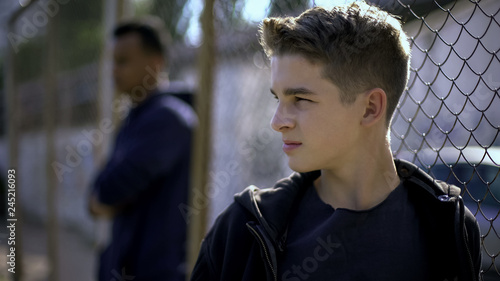 Thoughtful teen boys leaning on metal fence, upset with future, orphanage