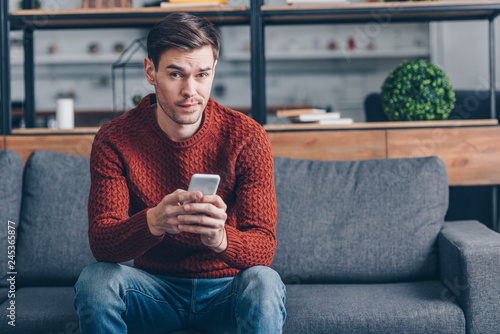 suspicious young man holding smartphone and looking at camera while sitting on couch at home