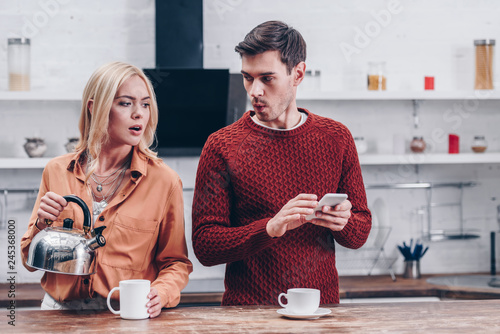 jealous young woman holding kettle and looking at husband with smartphone in kitchen