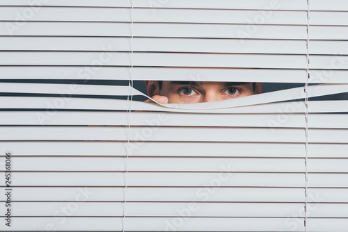 suspicious young man peeking and looking at camera through blinds, mistrust concept