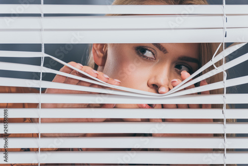 young woman looking away and peeking through blinds, mistrust concept