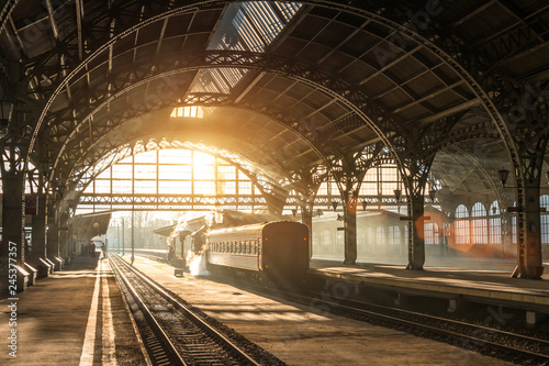Stary dworzec kolejowy z pociągiem i lokomotywą na peronie czekającym na odjazd. Wieczorne promienie słońca w łukach dymu.