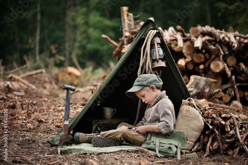 The boy, 5 years old, looks like a trapper, wanderer, lumberjack. Hut, shelter in the forest among logs of wood. A lonely walk, rest after work. Survival.
