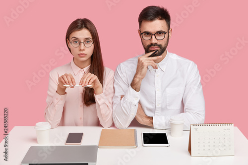 Joyful self assured woman with positive smile holds pen, serious bearded man in white shirt, collaborate together, use modern gadgets, pose at white table against pink background. Collaboration