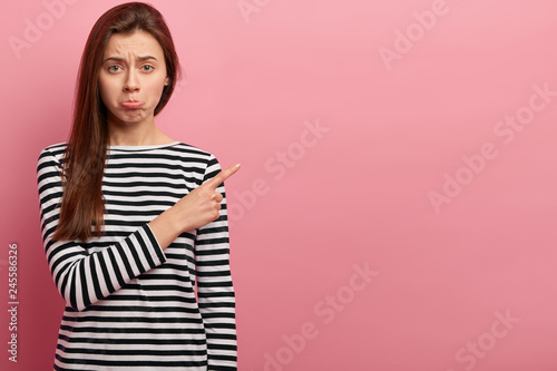 Unlucky gloomy European woman makes unhappy face, points aside with regret, feels sadness, wears casual striped clothes, stands against pink wall with blank space for your promotional content