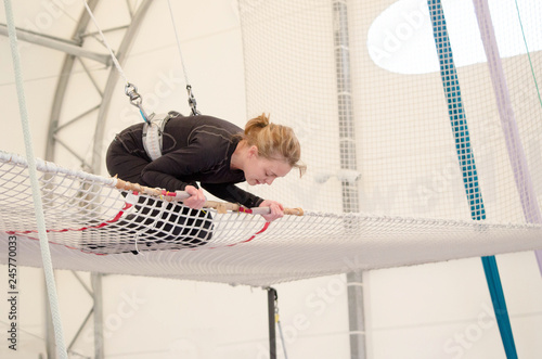 An adult female lands on a net, preparing to dismount at a on a flying trapeze school at an indoor gym. The woman is an amateur trapeze artist.