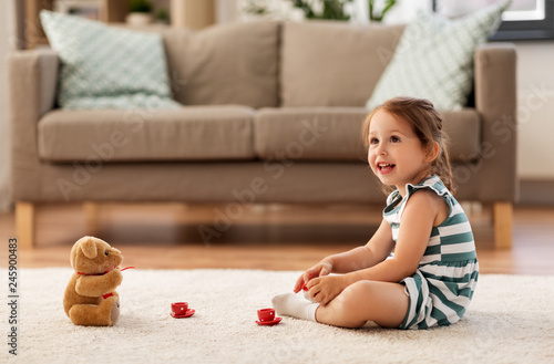 childhood and people concept - happy three years old baby girl playing tea party with toy crockery and teddy bear at home