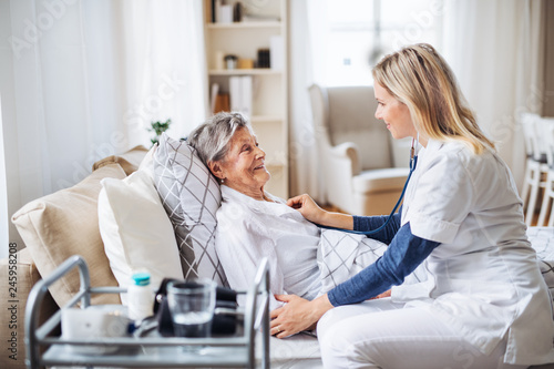 A health visitor examining a sick senior woman lying in bed at home with stethoscope.