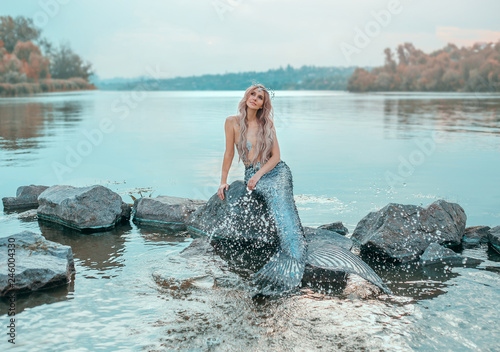 fair-haired mermaid in love dreams of handsome prince, new story Ariel, image of fairy-tale siren with long fish tail, chic crown of shells, plays, sprinkles clear water, ecology of environment