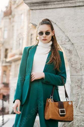 Outdoor fashion portrait of young beautiful fashionable woman wearing sunglasses, white turtleneck, green suit, blazer, trousers, holding brown suede bag, posing in street of european city 