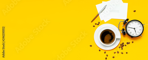Flat lay cup black coffee, coffee beans, black alarm clock, pens, white cards on yellow background top view copy space. Creative Concept time to work, female desktop, coffee background