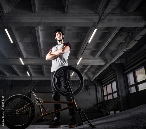 Full body portrait of a young man in protective helmet with his bike with his arms crossed standing in a skatepark indoors
