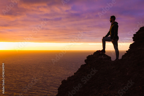 Visionary man standing on top of cliff edge staring at colorful sunset by the sea in Gran Canaria. Silhouette of person witnessing unique twilight from mountain top. Successful, entrepreneur concepts