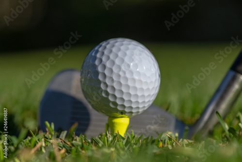 White golf ball teed up on a yellow tee with club face behind it and with soft green background
