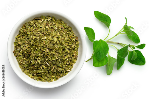 Dried chopped oregano in white ceramic bowl next to fresh oregano leaves isolated on white from above.