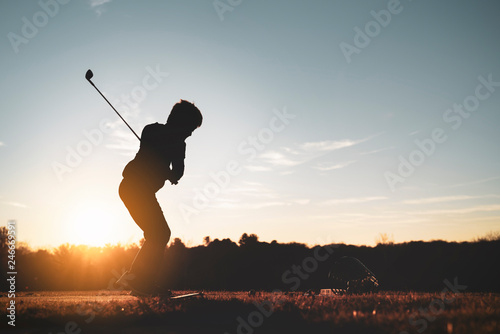Young junior golfer practicing in a driving range with beautiful sunset light in winter.