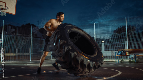 Strong Muscular Fit Young Shirtless Man is Doing Exercises in a Fenced Outdoor Basketball Court. He's Flipping a Big Heavy Tire in an Afternoon After Rain in a Residential Neighborhood Area.