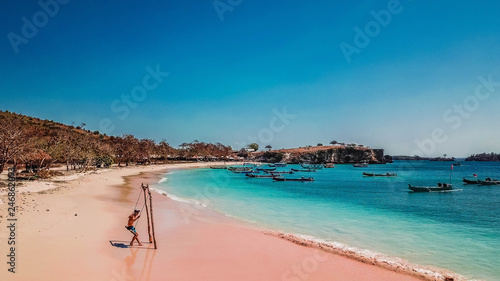 A man swinging on the Pink Beach in Lombook, Indonesia. Beautiful beach. Bay full of boats. No other people. Beautiful colors of the sand and the water. Dried trees in the background.