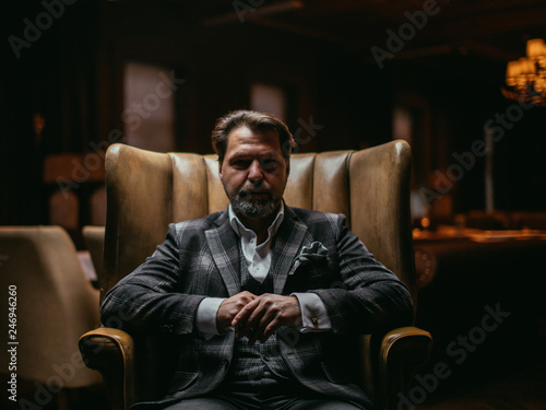 Pensive elegant bearded businessman thinks about something important while sitting in the leather armchair in living room