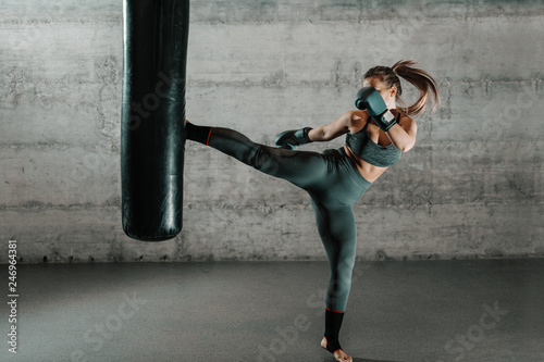 Caucasian woman in sportswear and with boxing gloves kicking bag in the gym. Full length. Wall in background.