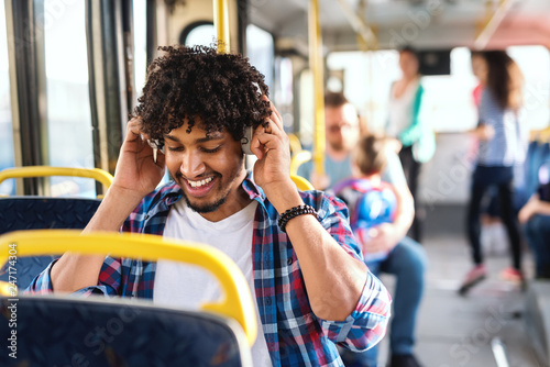 Smiling African guy sitting in the public transportation and putting on headphones. In background people sitting and standing.