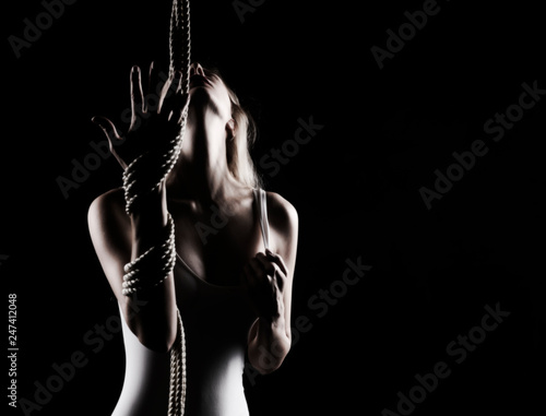Beautiful slender blonde girl with big breast and nipples appearing through clothes, wearing a white bodisuit, sensually plays with the ropes on black. Artistic noir silhouette photo