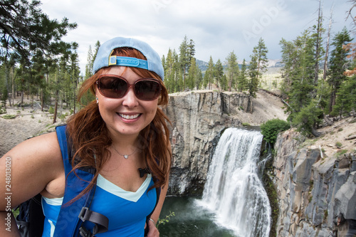 Redhead female hiker wearing a ballcap poses at Rainbow Falls in California along the John Muir Trail