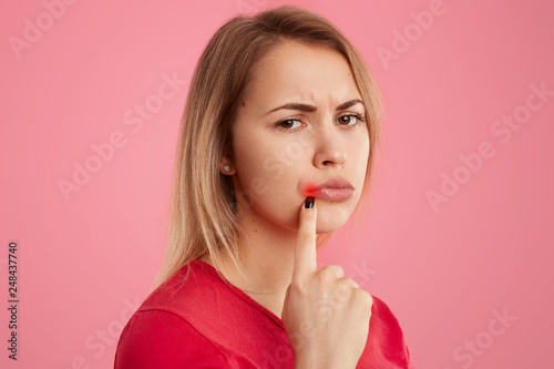 Close up shot of pretty woman being affected by herpes virus, keeps fore finger near lips, has dissatisfied facial expression, wears red clothing poses over pink background. People and illness concept