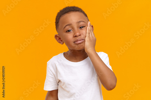 People, negative emotions, health and illness concept. Sad unhappy dark skinned male child having painful upset look, going to cry while suffering from intolerable toothache, touching chin