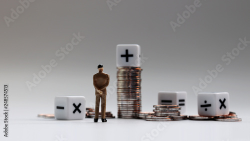 The back of a miniature businessman standing with a pile of coins and an arithmetic symbol cube.