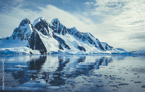 Ice covered mountains in polar ocean. Winter Antarctic landscape in blue and white tints. The mount's reflection in the crystal clear water. The cloudy sky over the massive glacier. Travel wild nature