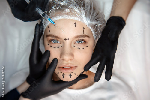 Calm peaceful young woman lying on couch and look straight forward. Her face is marked. Three hands touching face skin. Fourth hold syringe for injection.