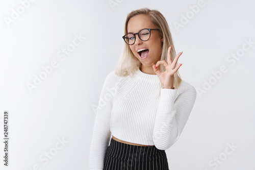 Emotive charismatic and happy young energized woman ready for work feeling uplifted, healthy after drinking vitamins showing okay gesture winking cheeky being in excellent mood over white background