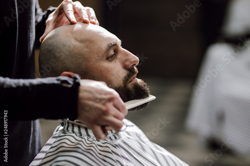 Fashion barber dressed in a black clothes tidies up men's beard  and scissors it in the barbershop