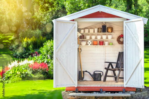 Garden shed filled with gardening tools. Shovels, rake, pots, water pitcher in storage hut. Green sunny garden in the background.