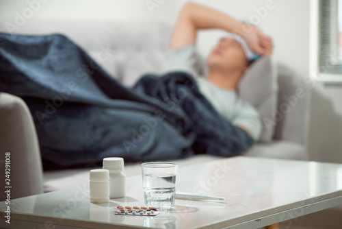 sick wasted man lying in sofa suffering cold and winter flu virus having medicine tablets in health care concept looking temperature on thermometer